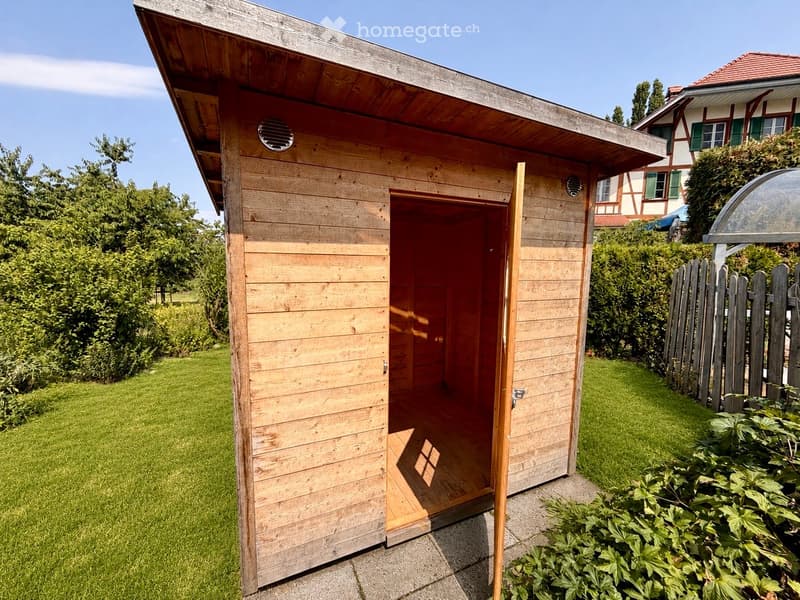 A wooden shed with an open door, situated in a well-maintained garden with green grass, bushes, and trees. The shed has a slanted wooden roof and is surrounded by a wooden fence and a traditional house in the background.