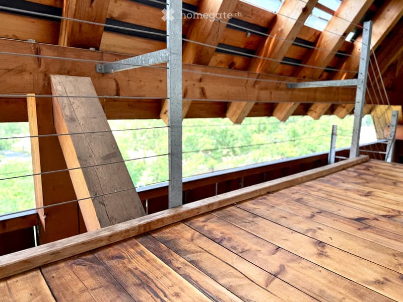 Wooden balcony with metal railings and exposed wooden beams, overlooking a green outdoor area.
