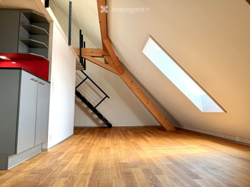 Empty attic space with wooden flooring, slanted ceilings, and a staircase leading upwards. Natural light streams in from a skylight window.