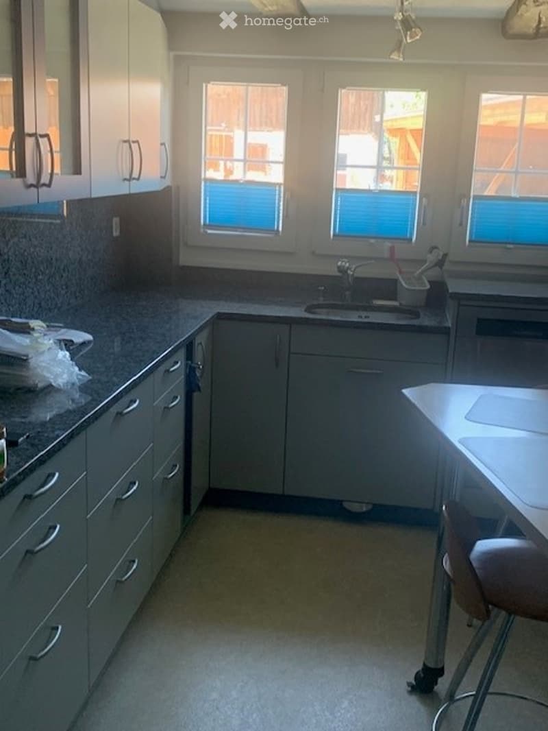 Modern kitchen with white cabinets, granite countertops, and stainless steel sink. Features three windows, a small table with a stool, and a few items on the counter.
