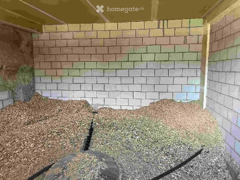 A small garden area with a raised wooden deck, surrounded by a brick wall. The ground is covered with mulch and small patches of grass, and there is a tree trunk visible on the left side.