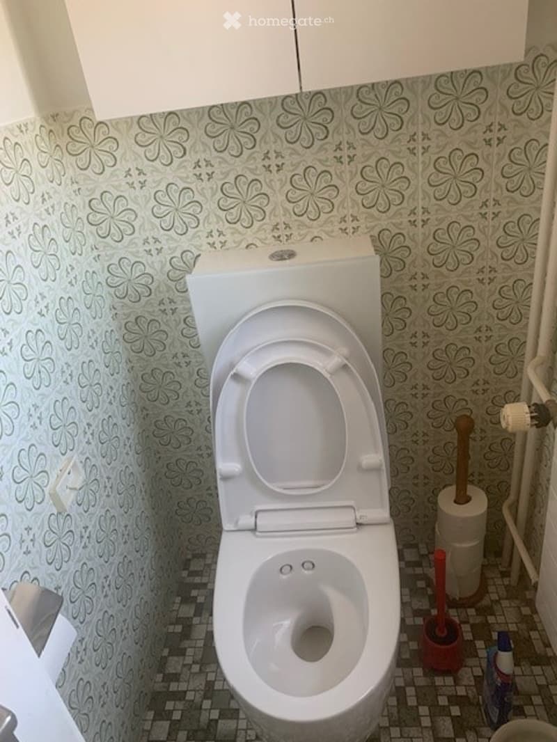 A small bathroom with a white toilet, floral-patterned wallpaper, and a tiled floor. Toilet paper and a cleaning spray bottle are visible.