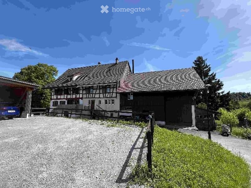 Traditional half-timbered house with a tiled roof, surrounded by a gravel driveway and a small garden area. A blue car is parked in the garage.