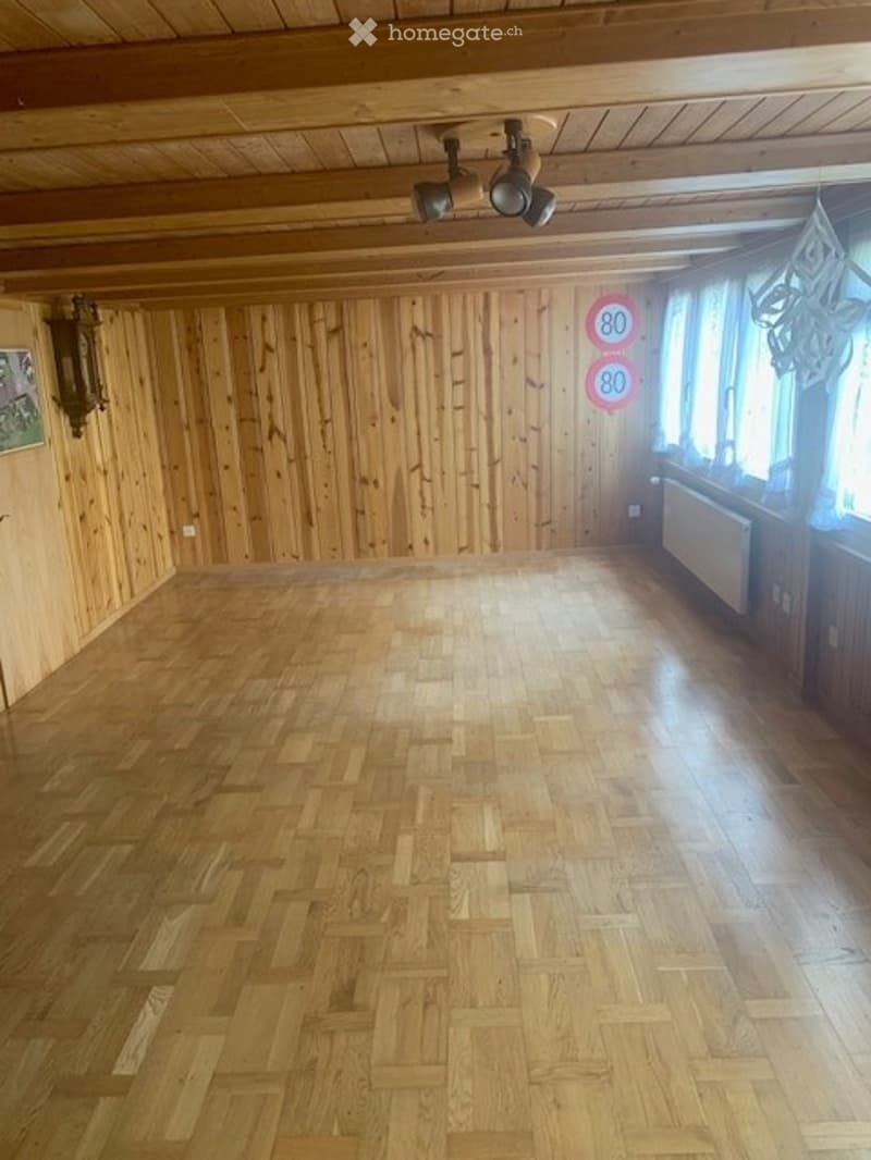 Completely empty room with wooden walls and herringbone wooden flooring, exposed wooden ceiling beams, and large windows with curtains.