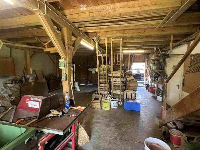 A spacious workshop with wooden beams and exposed ceiling, featuring a workbench, various tools, storage cabinets, and scattered items like buckets and boxes.