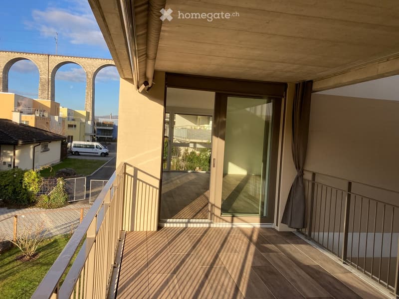 Balcony with wooden flooring, metal railing, large glass sliding doors, and a view of a residential area with a stone aqueduct in the background.