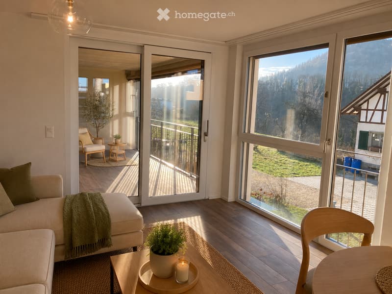 Bright living room with large windows and sliding doors leading to a balcony, featuring a sofa, coffee table, and a small table with a candle and potted plant.