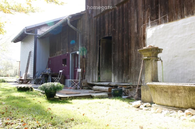 Rustic outdoor area with a wooden barn, a small patio with a table and chairs, a stone fountain, and a potted plant.