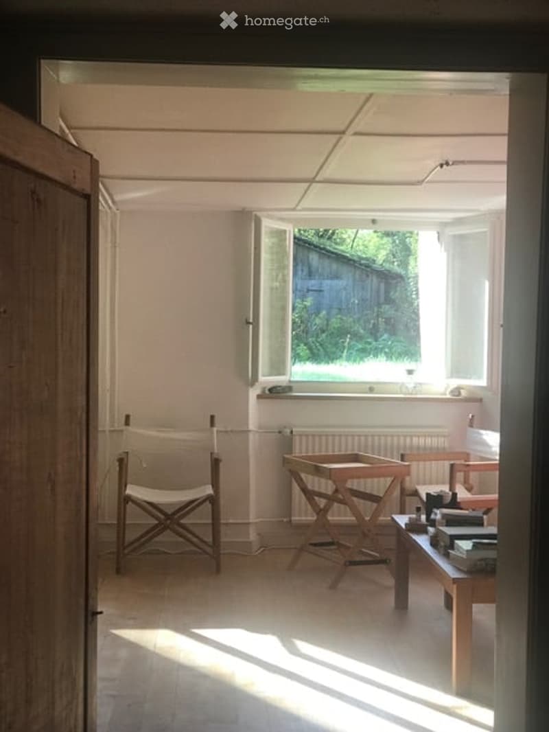 Bright, minimalist room with wooden flooring, white walls, and large windows. Contains a simple wooden chair, a small table, a stool, and a laptop on a surface.