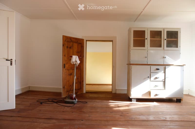 A room with wooden flooring, white walls, and a white cabinet with glass doors and drawers. A vacuum cleaner is placed on the floor near the door.