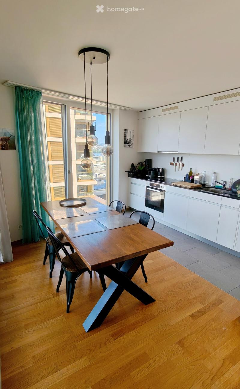 Modern kitchen with white cabinets, wooden dining table, chairs, and a view of the city through large windows.