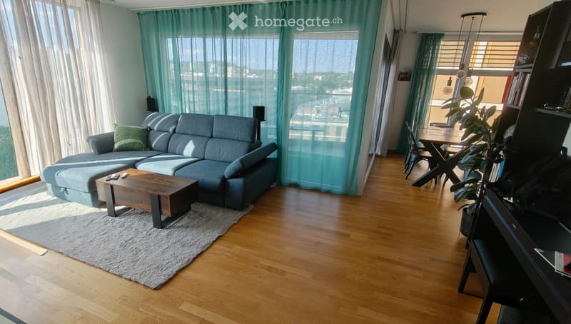Modern living room with a grey sectional sofa, wooden coffee table, dining area with chairs and table, bookshelf, and a potted plant. Large windows with sheer curtains let in natural light.