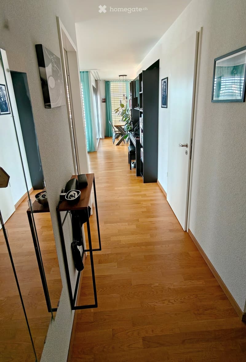 A long, narrow hallway with wooden flooring, white walls, and several doors leading to different rooms. There are framed pictures on the walls and a small table with items on it to the left.