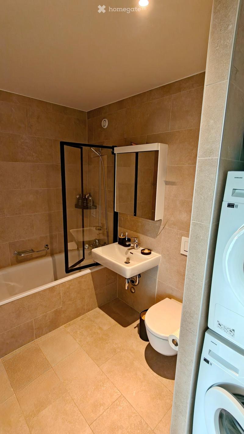 Modern bathroom with beige tiles, a white sink, toilet, bathtub, and a glass-enclosed shower. A washing machine is visible on the right side.