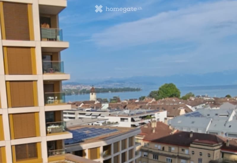 View from a balcony showing a cityscape with rooftops, a lake, and distant mountains.