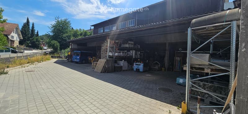 A large industrial-style building with an open garage area containing various construction materials, tools, and equipment. The paved area in front is clean and organized.