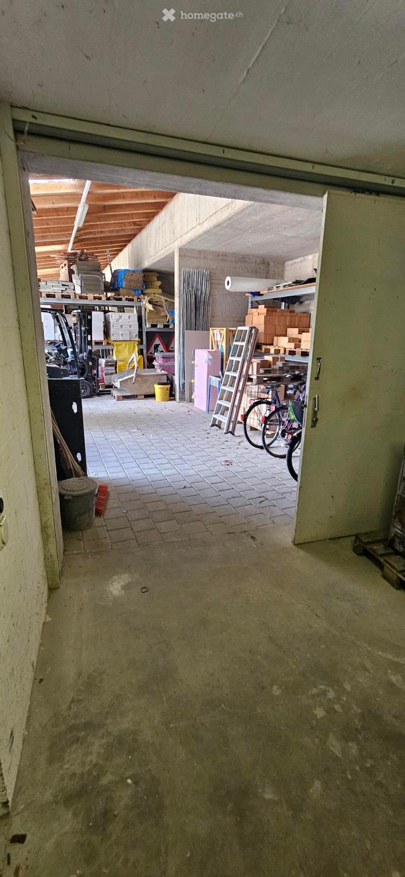 Empty room with unfinished concrete walls and floor, leading to a storage area with boxes, tools, and bicycles.
