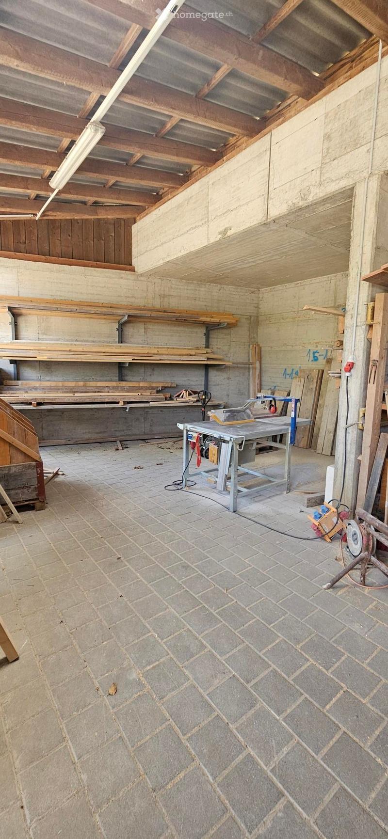 Empty workshop with exposed wooden beams, concrete walls, tiled floor, and wooden shelves holding planks and tools.