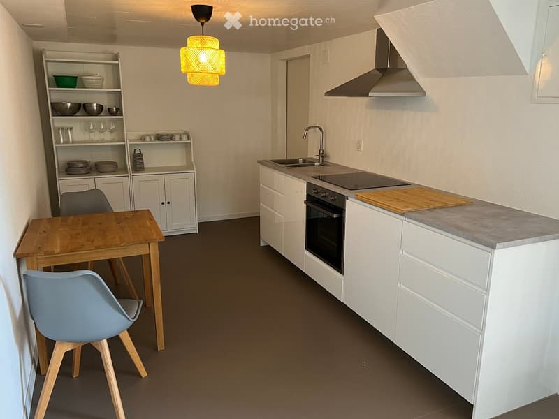 Modern kitchen with white cabinets, grey countertops, a table, and a wooden chair. Oven, stove, and a sink with a faucet. A hanging lamp is present.