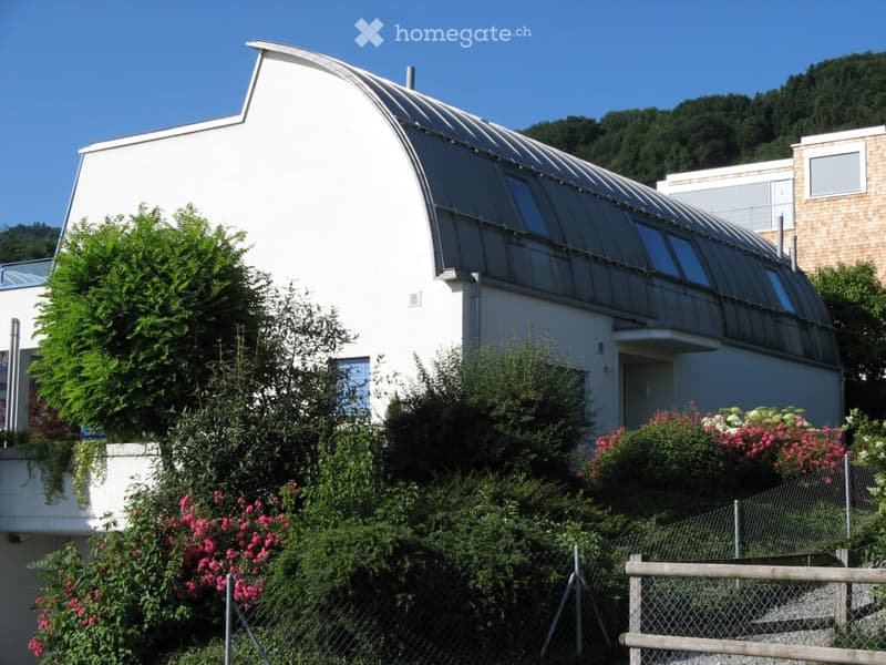 Modern white building with a curved roof and large glass panels, surrounded by lush greenery and flowering plants.