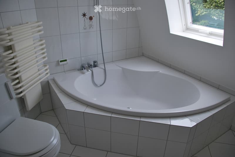 Modern bathroom with a large, curved bathtub, toilet, radiator, and a window letting in natural light.
