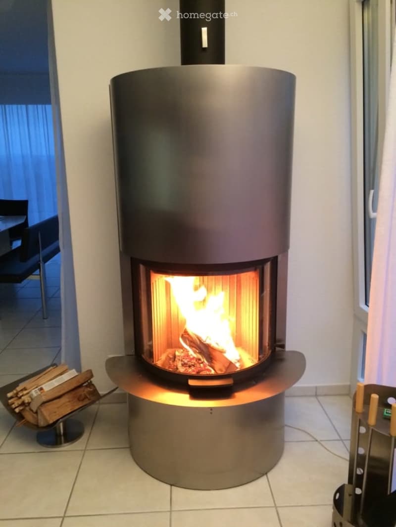 Modern, sleek black cylindrical fireplace with a glowing fire inside, surrounded by a polished metal base. A small stack of firewood is placed nearby, and a chair is visible in the background.