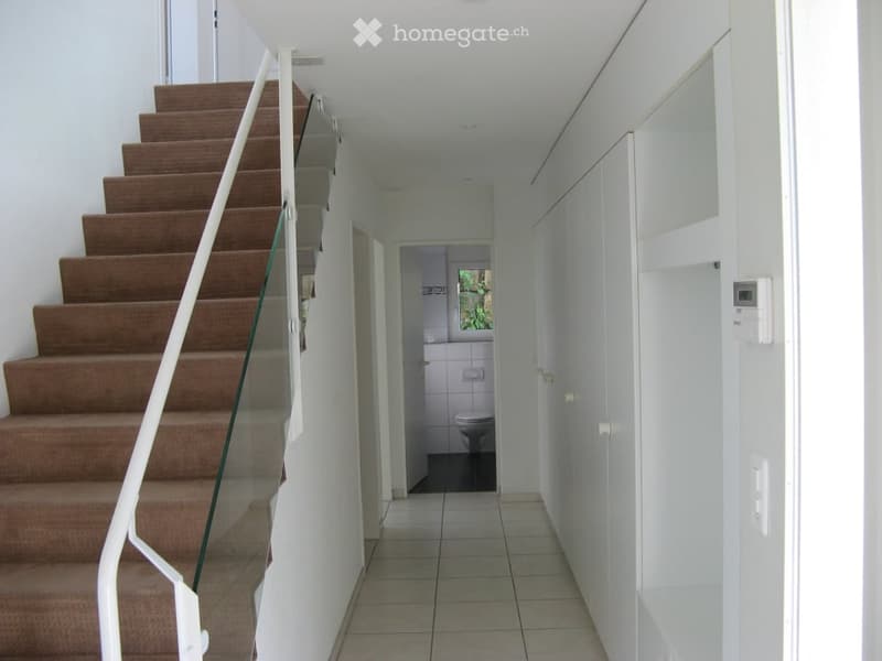 A narrow hallway with tiled flooring, white walls, and a staircase leading upwards. A small bathroom is visible at the end of the hallway.