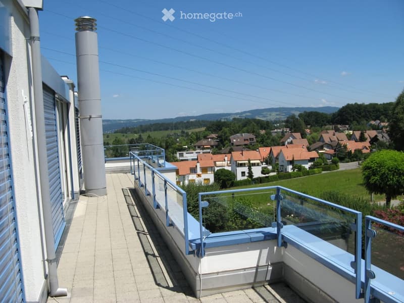 Balcony with a view of a residential area and distant hills, featuring a glass railing and tiled floor.