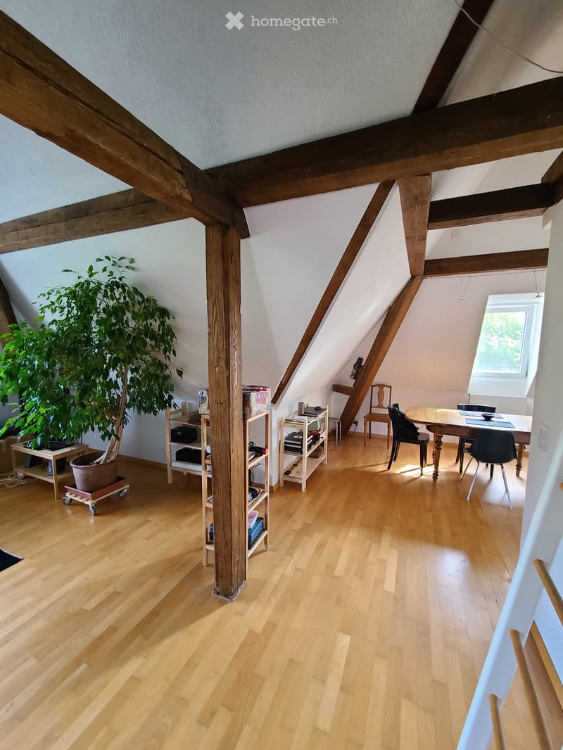 Bright attic room with wooden floors, slanted wooden ceiling beams, large window, and modern furniture including a dining table, chairs, shelves, and a potted plant.