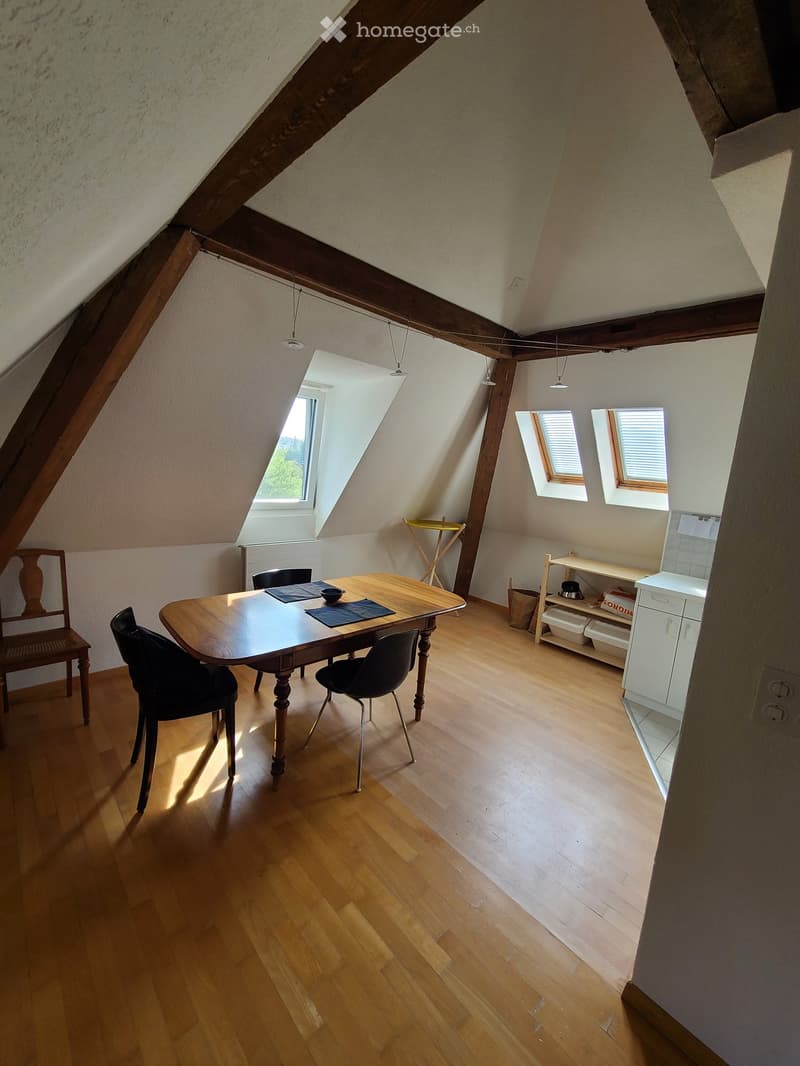 Bright attic room with wooden floors, slanted wooden ceiling beams, and multiple windows. Contains a dining table with chairs, a small shelf, and a white cabinet.