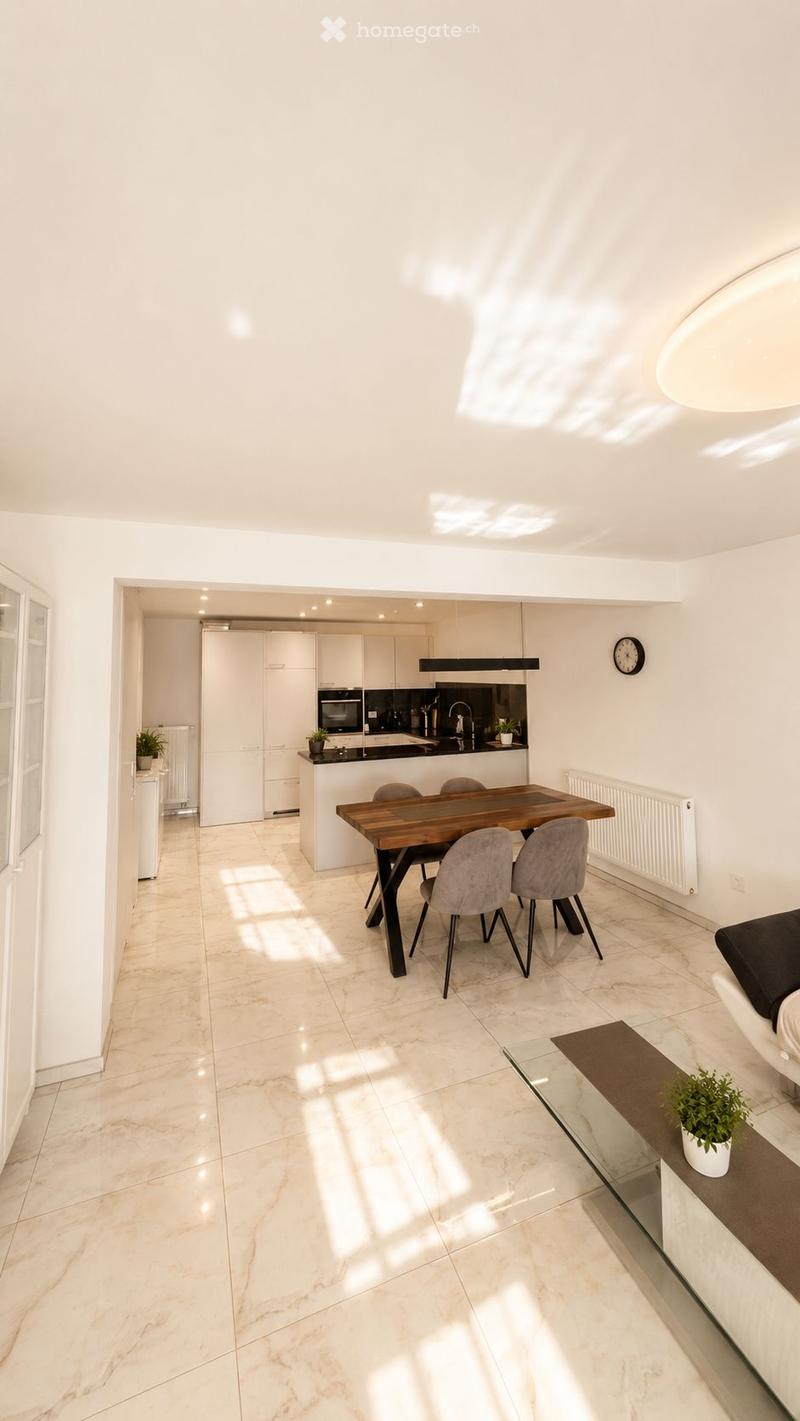 Bright, open-plan living and dining area with white walls, marble flooring, and modern furniture including a wooden dining table, grey chairs, a sofa, and a small kitchen area with white cabinets and stainless steel appliances.