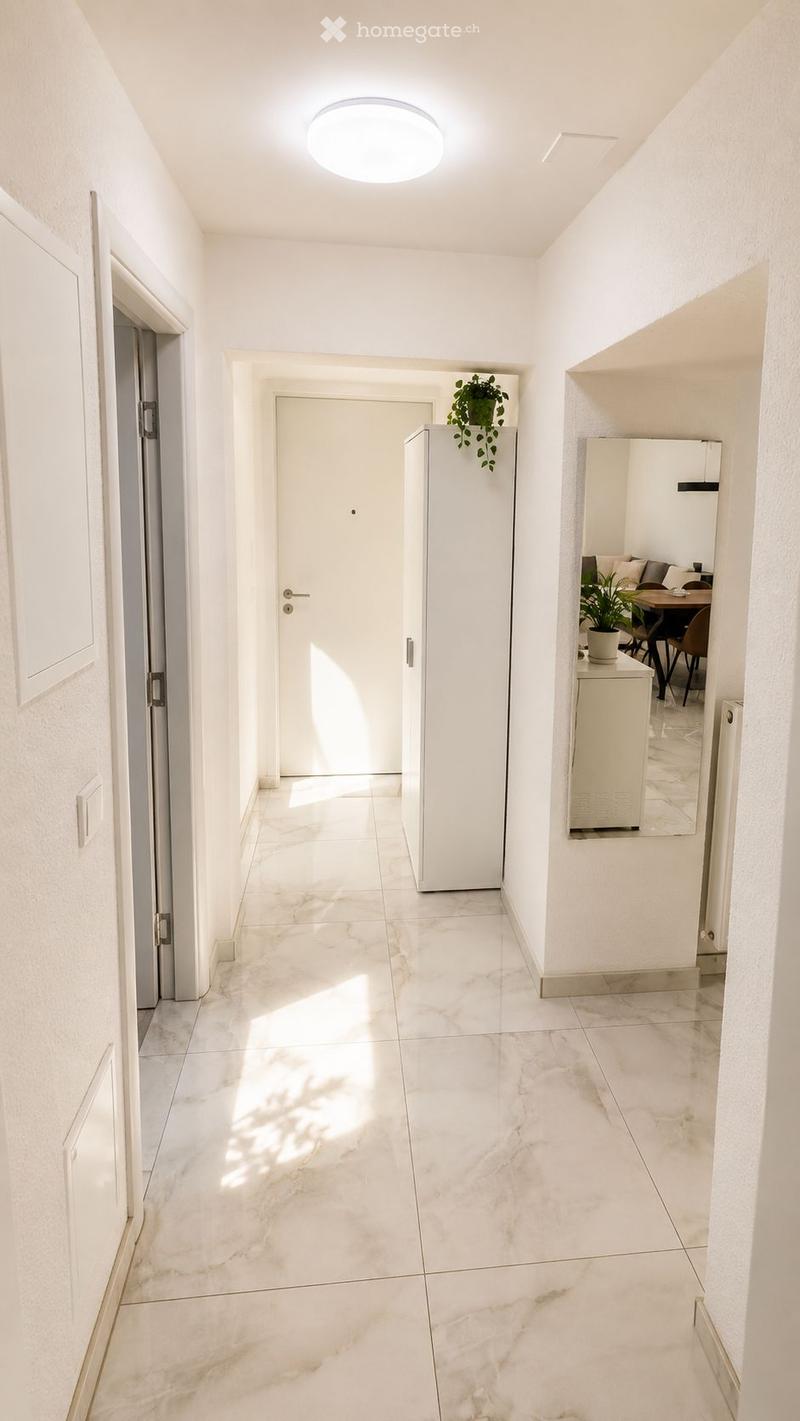 Bright, clean hallway with white walls, marble flooring, and recessed lighting. Features a closed door, a small plant on a shelf, and a glimpse of a furnished room with a sofa and dining table.