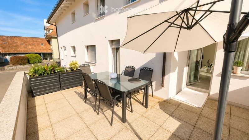 Balcony with a glass table, four chairs, and a large umbrella. Potted plants and a view of the house exterior.