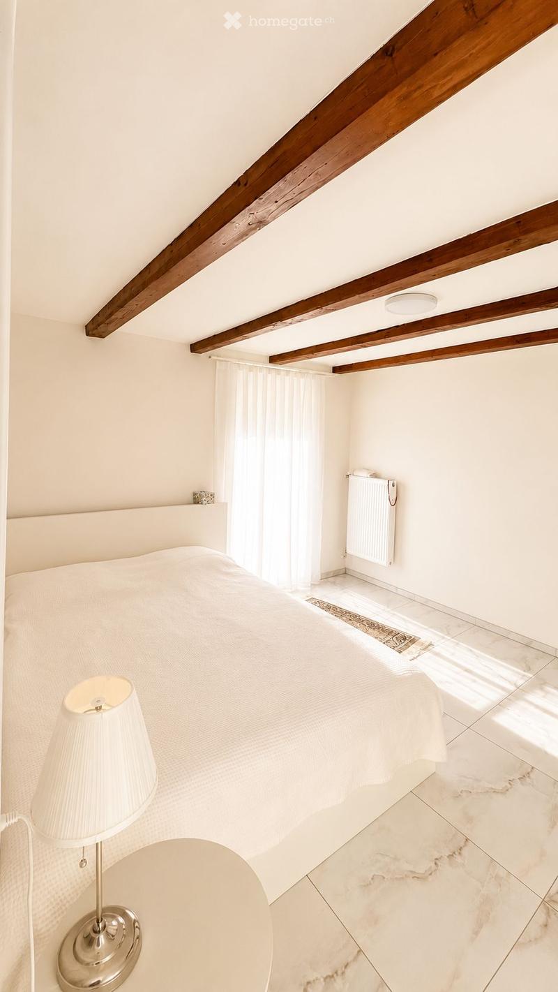 Bright bedroom with a large bed, a bedside table with a lamp, exposed wooden beams on the ceiling, and a radiator. The room features marble flooring and large windows with white curtains.