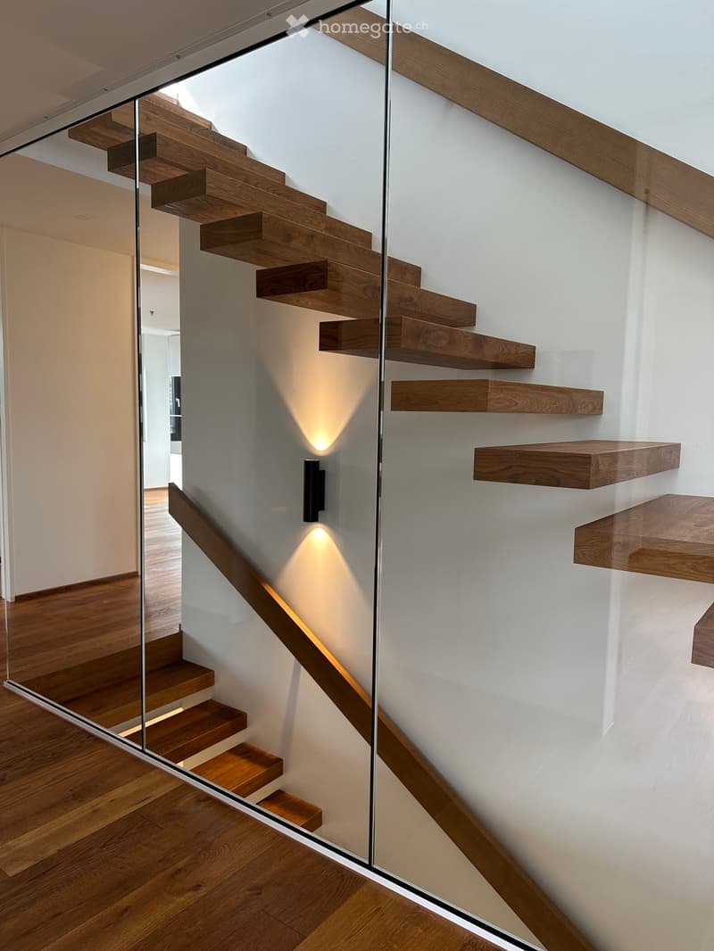 Modern staircase with wooden steps and a glass railing, illuminated by wall-mounted spotlights, leading to an open area with wooden flooring.