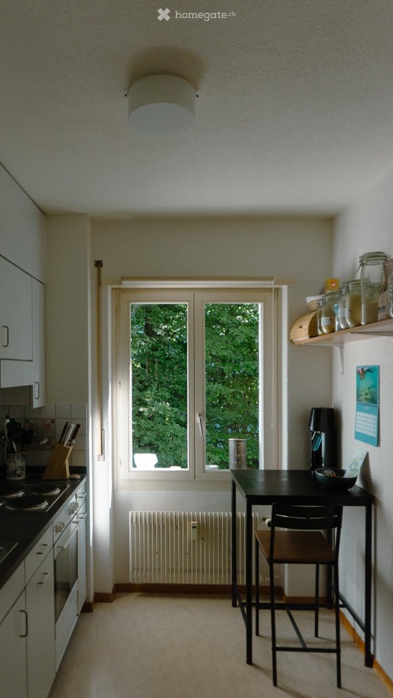 Bright kitchen with white cabinets, a black dining table and chair, a stove, and shelves with jars and a calendar.