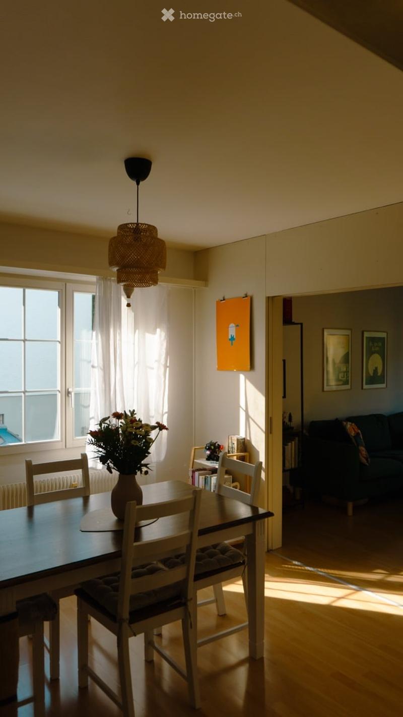 Bright dining area with wooden floor, large windows, dining table with chairs, vase with flowers, pendant lamp, and adjacent living space with sofa.