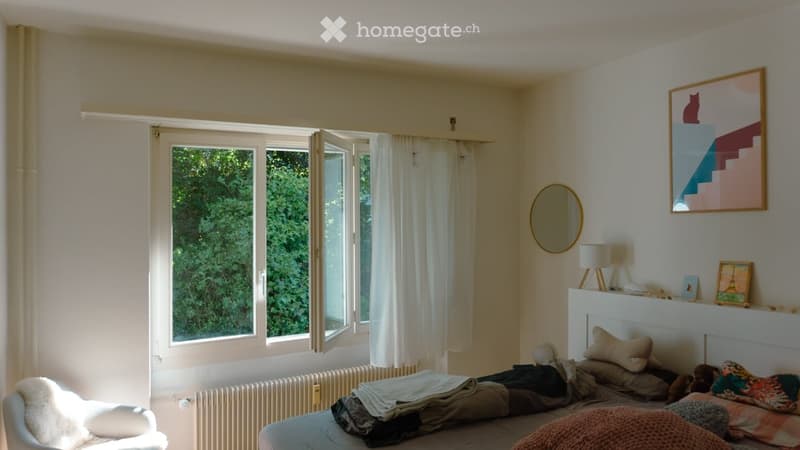 Cozy bedroom with a bed, a white chair, a round mirror, a small table lamp, and a colorful picture frame on the wall. Large windows with white curtains let in natural light.