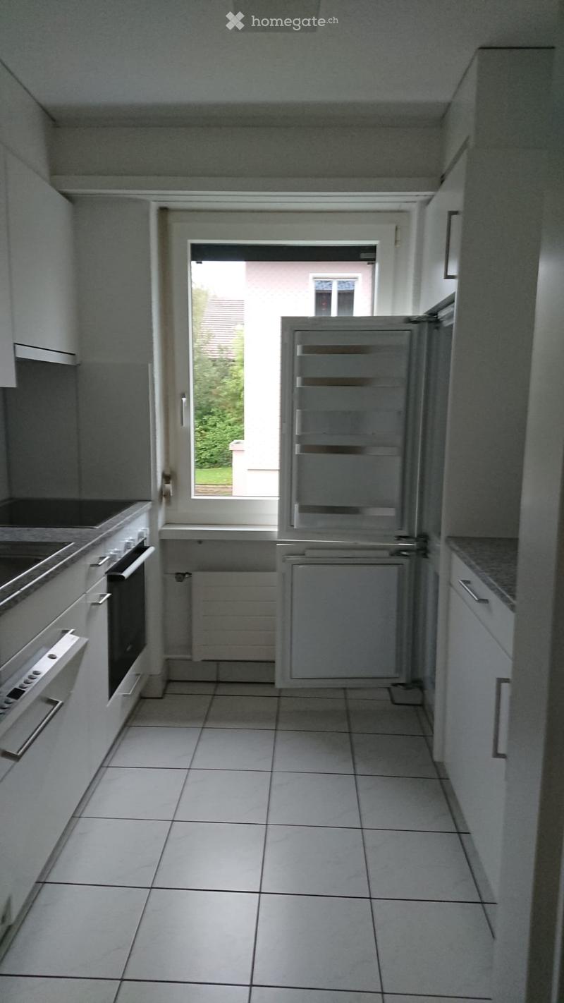 Modern kitchen with white tiled floor, white cabinets, and stainless steel appliances including a refrigerator and oven. A window provides natural light and a view of the outdoor area.