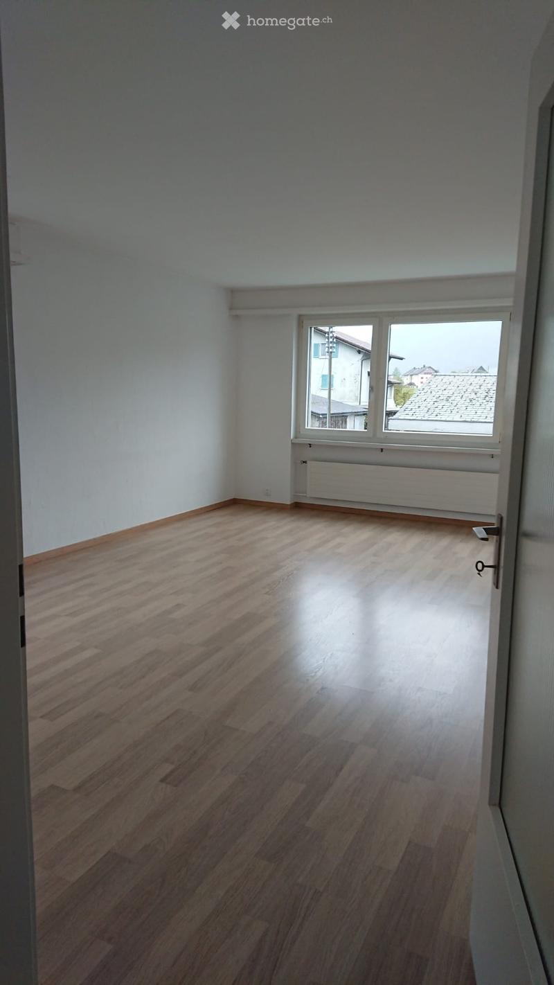 Empty room with wooden flooring, white walls, and a large window with a view of rooftops and buildings.
