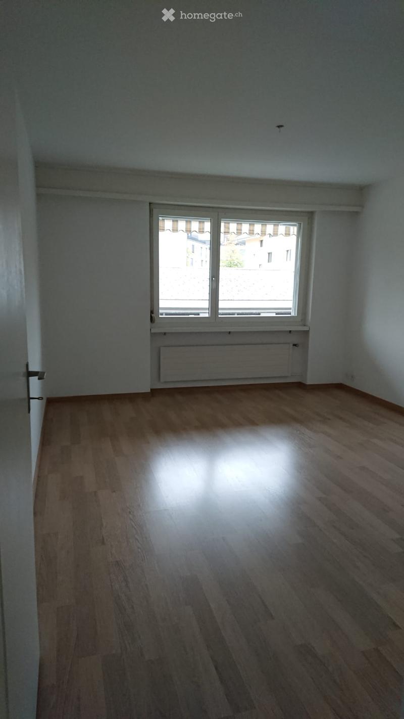 Empty room with wooden flooring, white walls, and a large window with blinds. A radiator is visible below the window.