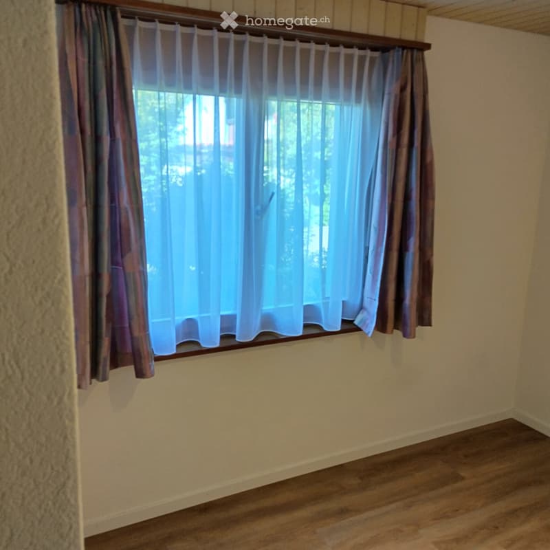 Empty room with wooden flooring, light-colored walls, and a large window with curtains.