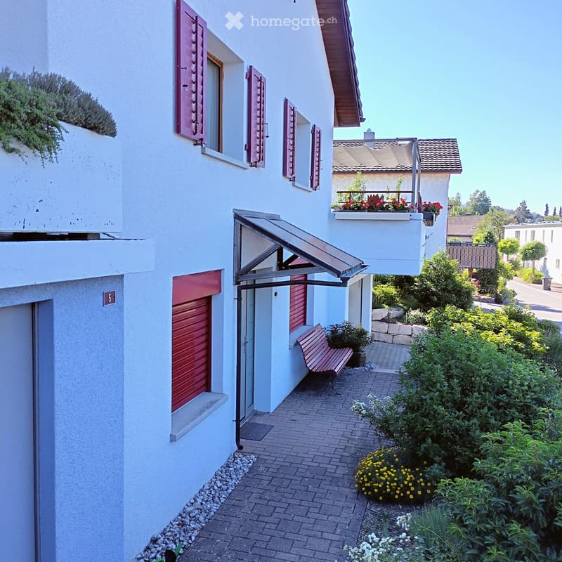 Two-story white building with red shutters, a covered entrance, and a small garden area with a bench and flowers.
