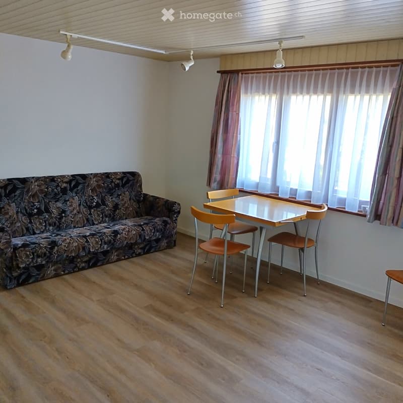 A furnished room with a sofa, a small table, and chairs, featuring wooden flooring and large windows with curtains.