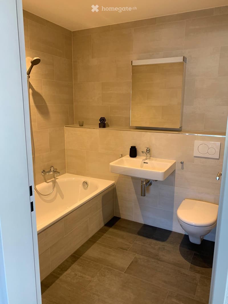Modern bathroom with beige tiles, a bathtub, a sink, a toilet, a mirror, and a showerhead.
