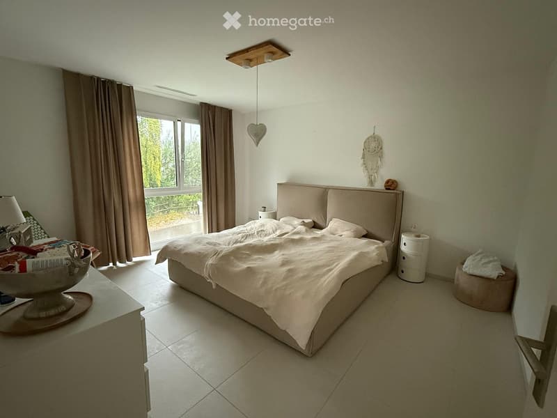 A modern bedroom with a large bed, nightstand, lamp, and a window with curtains. The room features a dreamcatcher on the wall and a decorative bowl on the nightstand.