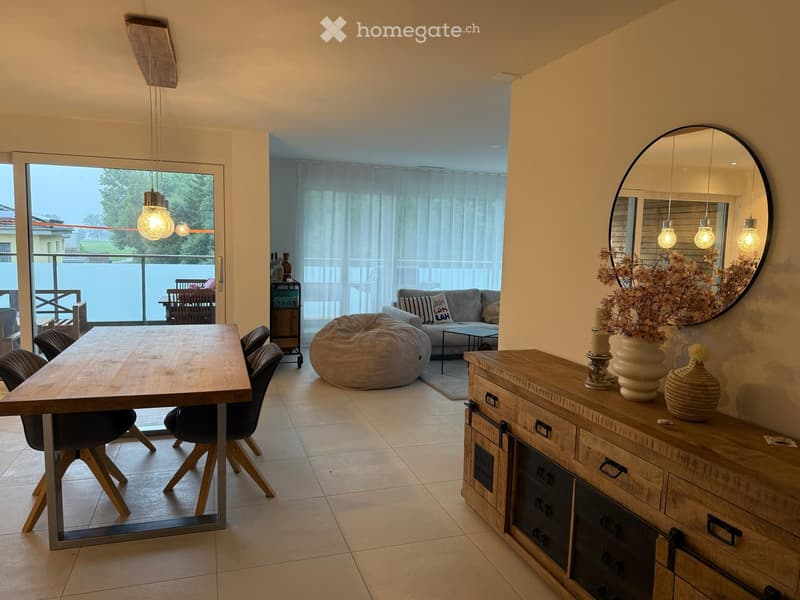 Bright living and dining area with a wooden dining table, chairs, sofa, coffee table, bean bag, large mirror, and decorative items on a wooden sideboard. Large windows and sliding doors lead to a balcony.
