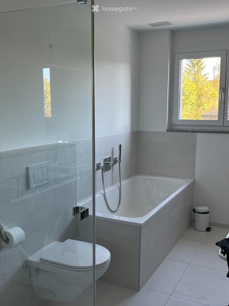 Modern bathroom with a bathtub, toilet, glass-enclosed shower, and a window offering a view of greenery.