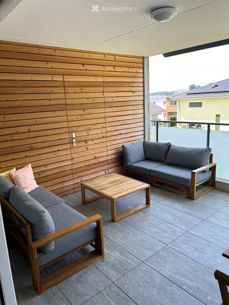 Balcony with grey sofas, wooden coffee table, and cushions, overlooking a suburban area with houses and solar panels.
