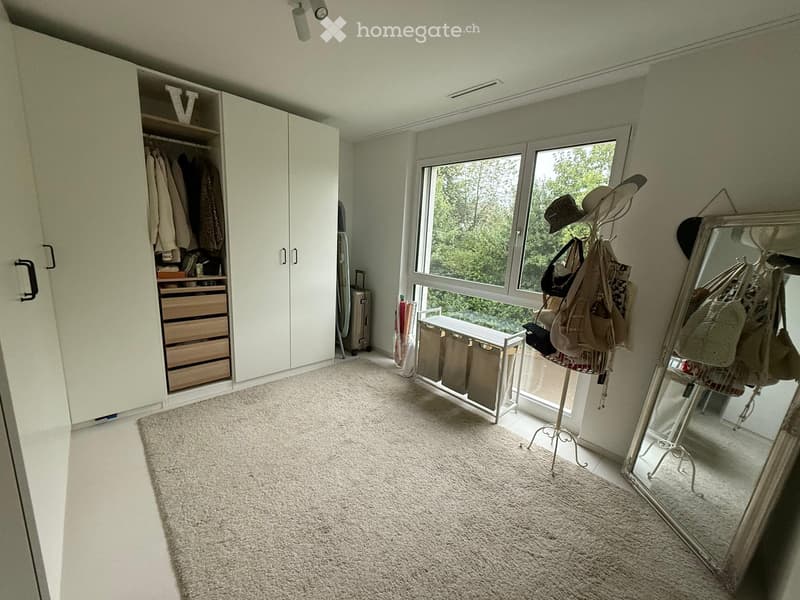 Well-lit room with a large window, white wardrobe with drawers, a bench, a mirror, and a hat rack with bags and hats.
